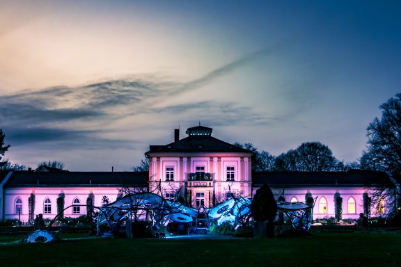 Historisches Gebäude mit bunten Lichtern, umgeben von Bäumen und einem klaren Himmel.