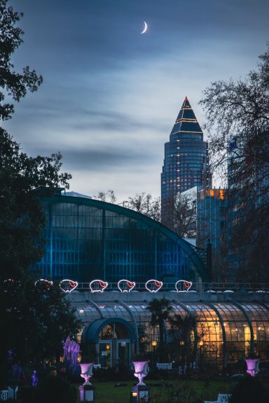Abendliche Stadtansicht mit Glasgewächshaus und Mond über einem modernen Hochhaus.