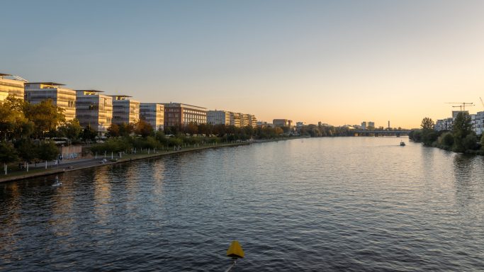 Stadtansicht am Wasser mit modernen Gebäuden und ruhiger Wasseroberfläche bei Sonnenuntergang.