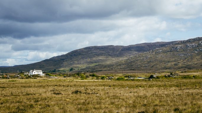 Landschaft mit hügeligem Gelände, einer Wiese und einem weißen Haus im Vordergrund.